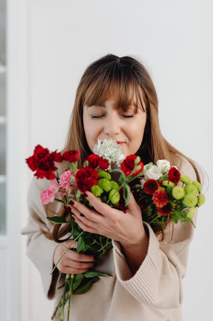 A young woman with long, light brown hair and bangs is holding a large bouquet of fresh flowers close to her face, eyes gently closed and a tender expression. The bouquet features vibrant red carnatio