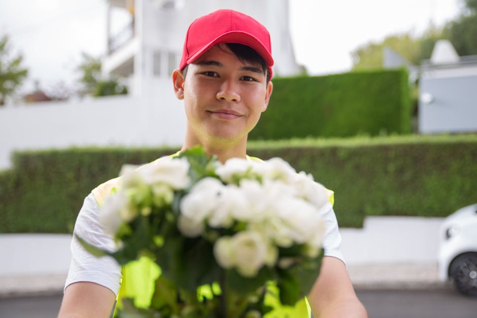 A delivery person wearing a red cap and a high-visibility vest holds out a bouquet of fresh, white roses and green foliage. The bouquet is arranged in a compact, rounded style, wrapped with clear plas