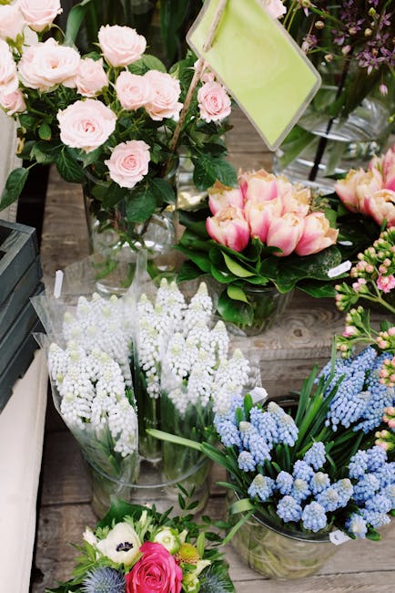 A vibrant display of fresh floral arrangements featuring pink roses in a clear glass vase, surrounding a bouquet of creamy pink tulips with green leaves, and clusters of white and light blue grape hya
