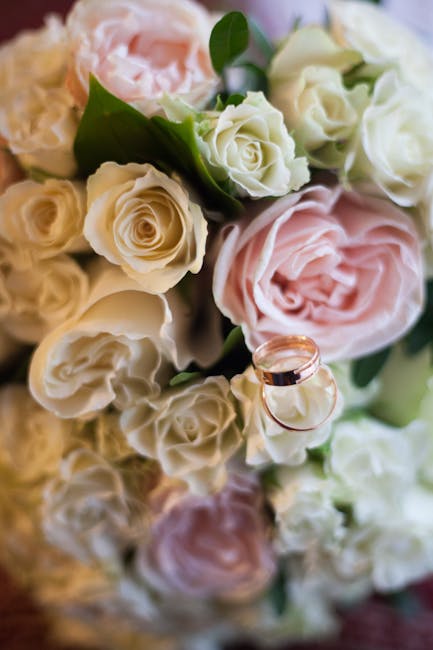 A close-up of a floral bouquet featuring soft pink and white roses, with some smaller white and cream-colored blooms, and lush green foliage. The roses display delicate, layered petals with a slightly