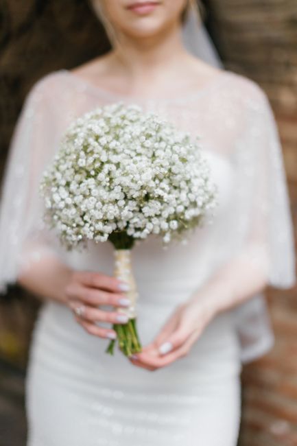 A bride holding a bouquet of delicate white baby's breath flowers with tiny, clustered blossoms and slender green stems wrapped in elegant cream ribbon. The bouquet contrasts softly with the bride's l