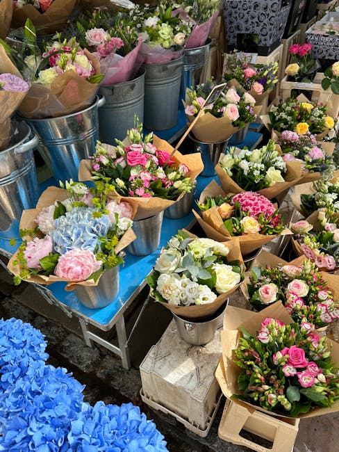 A variety of freshly arranged flower bouquets displayed on a table, featuring pastel pinks, whites, and soft blues, wrapped in brown paper or placed in metallic buckets and vases. The arrangements inc