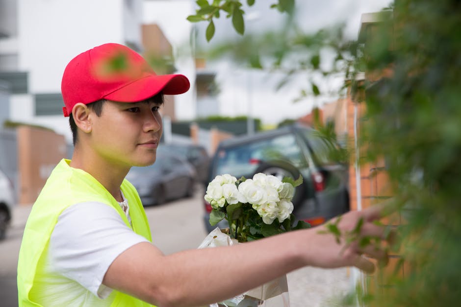 A delivery person wearing a red cap and a high-visibility yellow vest is placing a bouquet of fresh white flowers, likely roses or peonies, wrapped in light-colored paper, into a green leafy shrub out