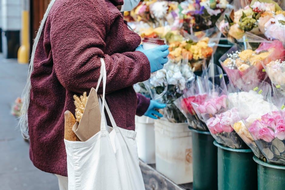A person wearing a maroon, textured fleece jacket and blue gloves is standing outdoors at a flower stall on Reigate High Street, holding a paper cup. They carry a white tote bag with French bread and 