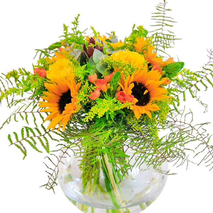 With its sun-drenched palette of sunflowers and yellow garden roses, this bouquet is a tangible expression of joy, arranged by skilled Reigate florists. The sunflower heads are expansive and brilliantly hued, their dark, textured centres ringed by petals that recall the brilliant yellows found along the River Mole in late summer. Nestled close, garden roses in creamy yellow spiral outward in tight formation-a nod to classic garden romance. Striking contrasts are provided by tight clusters of bright red hypericum berries sprinkled throughout, their gloss adding energy among the soft, matte blooms. A mix of lush greenery infuses the arrangement with fresh vitality: feathery ferns cascade at the rim, while sprigs of solidago and slender spikes stretch upward, echoing the wild beauty of local Reigate greenways. Subtle eucalyptus foliage threads throughout, adding a hint of silvery blue and a delicate, aromatic note reminiscent of early morning air in Memorial Park. The round glass vase, nearly spherical, holds the bouquet's stems submerged and glowing in clear water, grounding the composition and showcasing the artistry behind it. The overall effect is bright, joyful, and welcoming-perfect for birthdays, housewarmings, or to simply spread a spark of happiness. A testament to local expertise, this bouquet embodies Reigate's sense of occasion and its undeniable love of natural beauty.